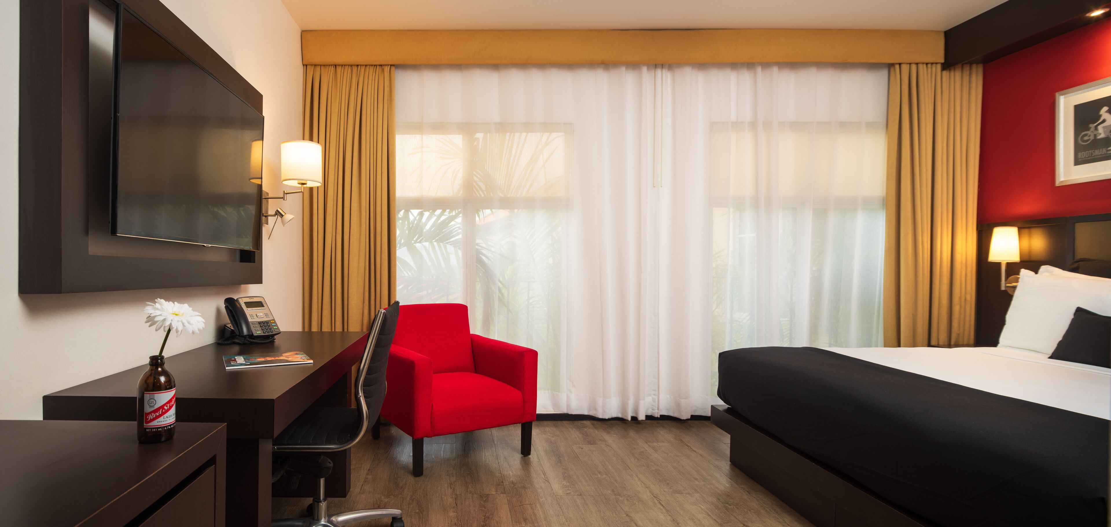 A hotel room with a bed, a desk with a chair, a TV mounted on the wall, and a window with curtains.