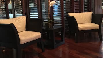 A room with a wooden floor, two dark armchairs with white cushions, a small table with a vase of flowers, and a window with wooden blinds.