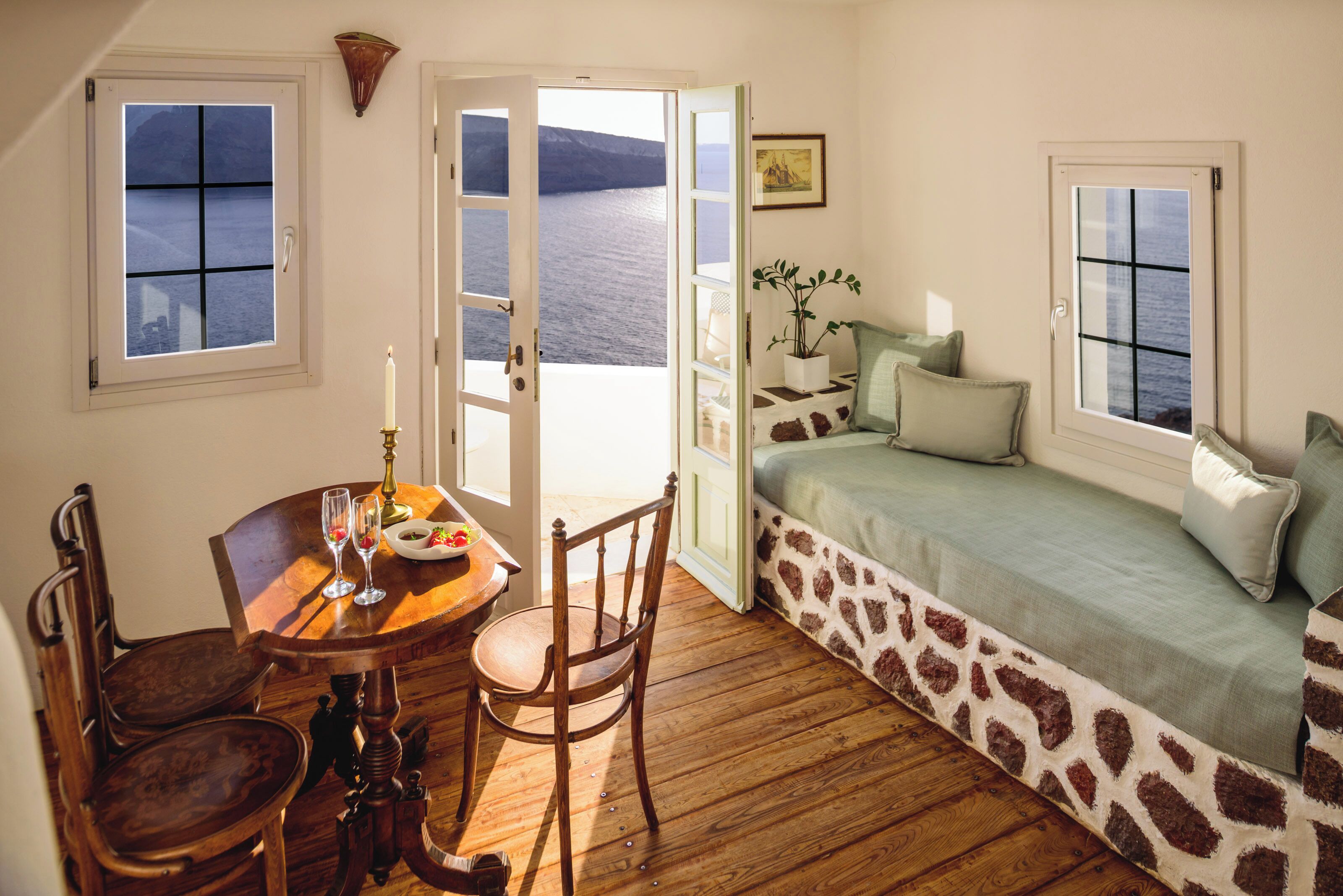 Une pièce dotée d’un plancher en bois, d’un lit recouvert d’une couvre-lit à motifs, d’une table à manger avec des chaises, et offrant une vue sur la mer par de grandes fenêtres ouvertes.