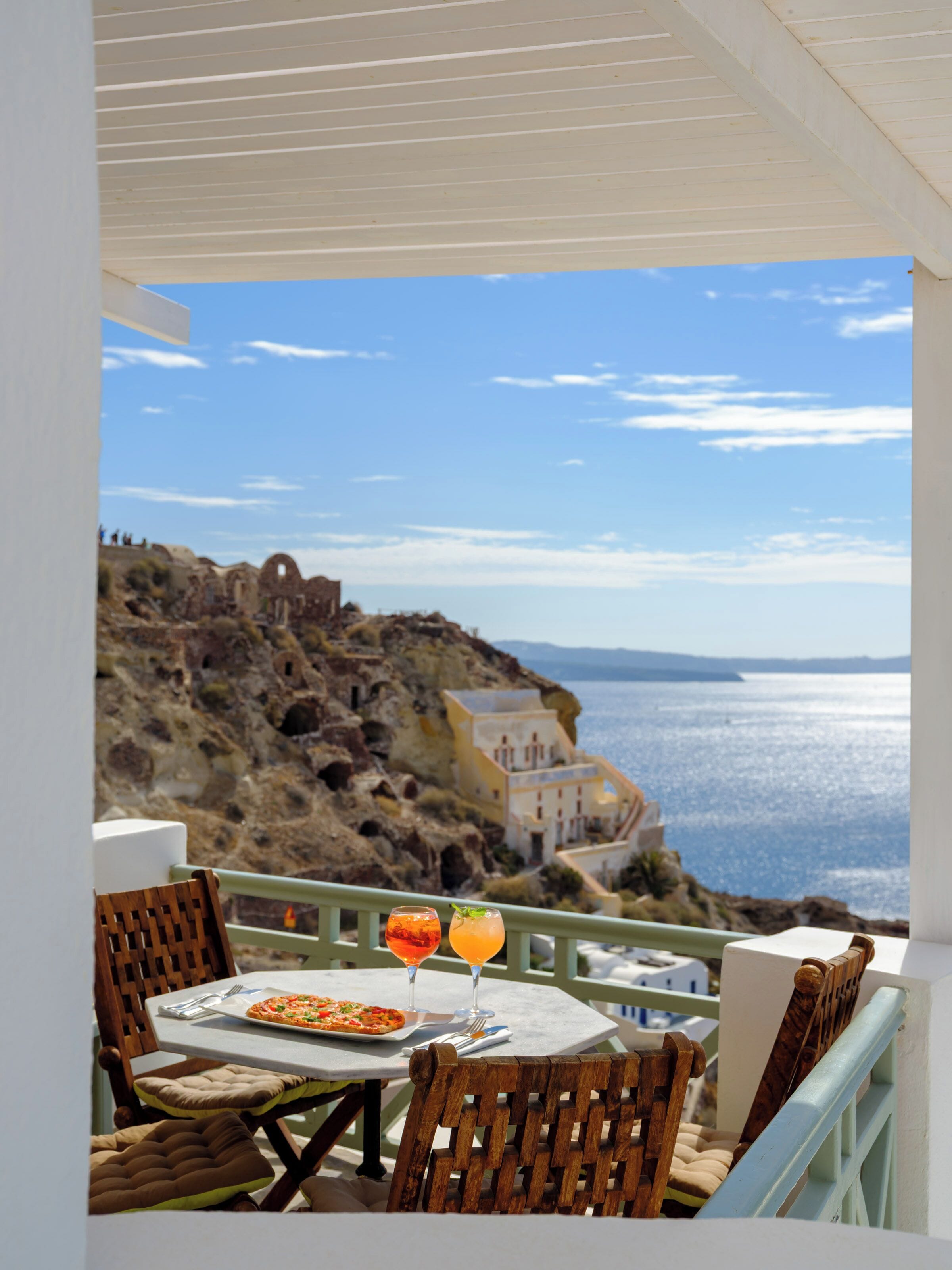 Una terraza con una mesa preparada para dos, con vista a un paisaje costero con ruinas y al mar.