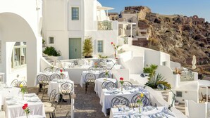 A white building with outdoor seating and tables, a rocky hillside in the background, and a clear blue sky. - Fanari Villas Santorini (Santorini)
