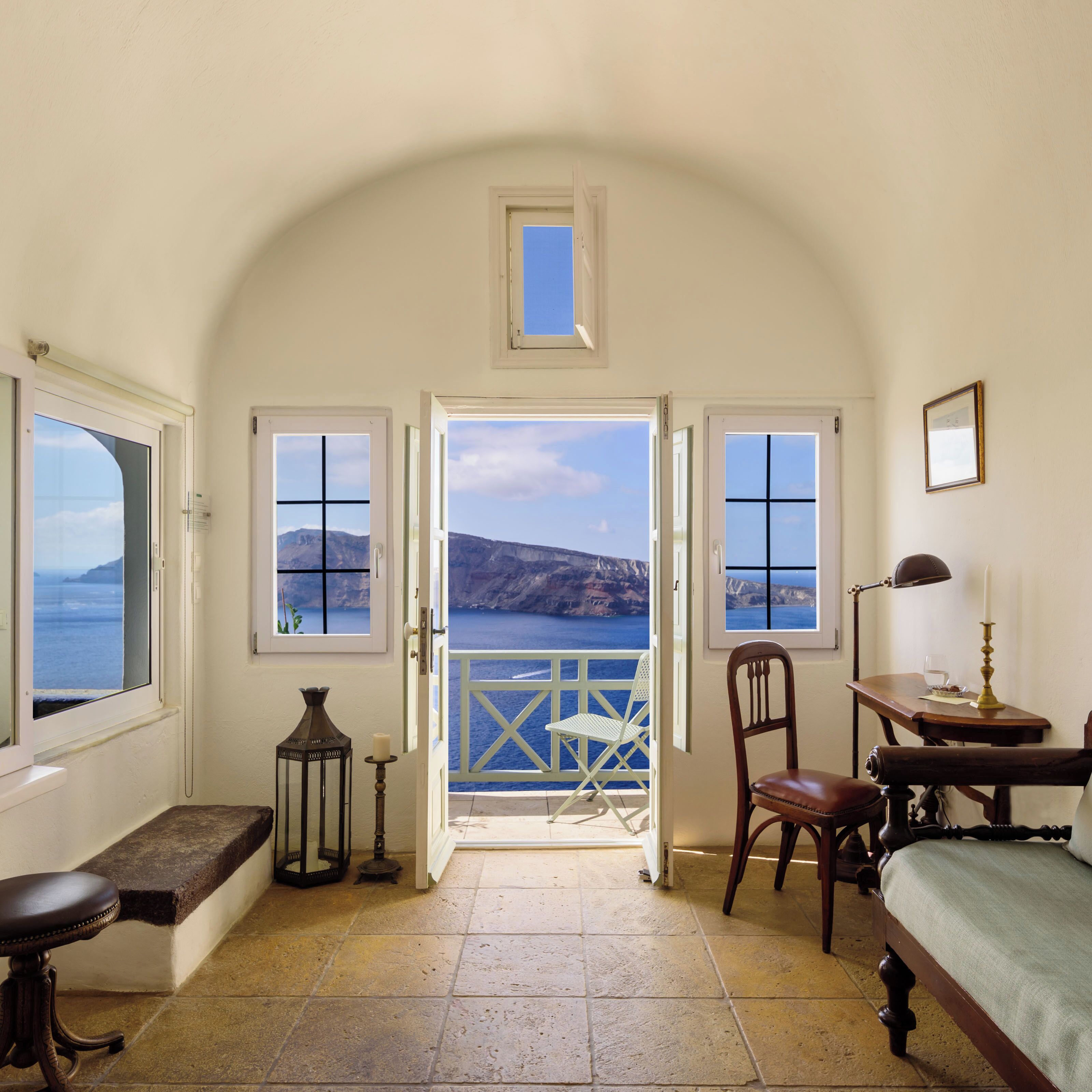 A room with a view of the sea, a wooden chair, a table with a lamp, and a bed.