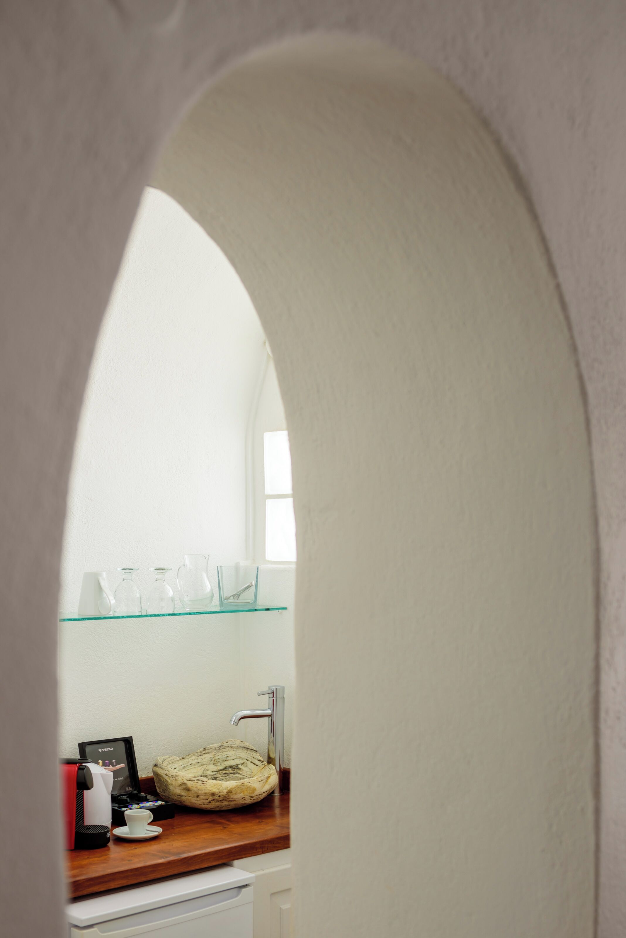 Vista a través de un arco de entrada a una habitación con repisas de vidrio que sostienen diversos objetos, una cafetera y una cesta.