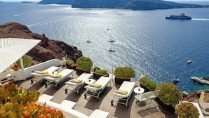 A deck with sun loungers and potted plants overlooking a calm sea with boats and a cruise ship. - Fanari Villas Santorini (Santorini)