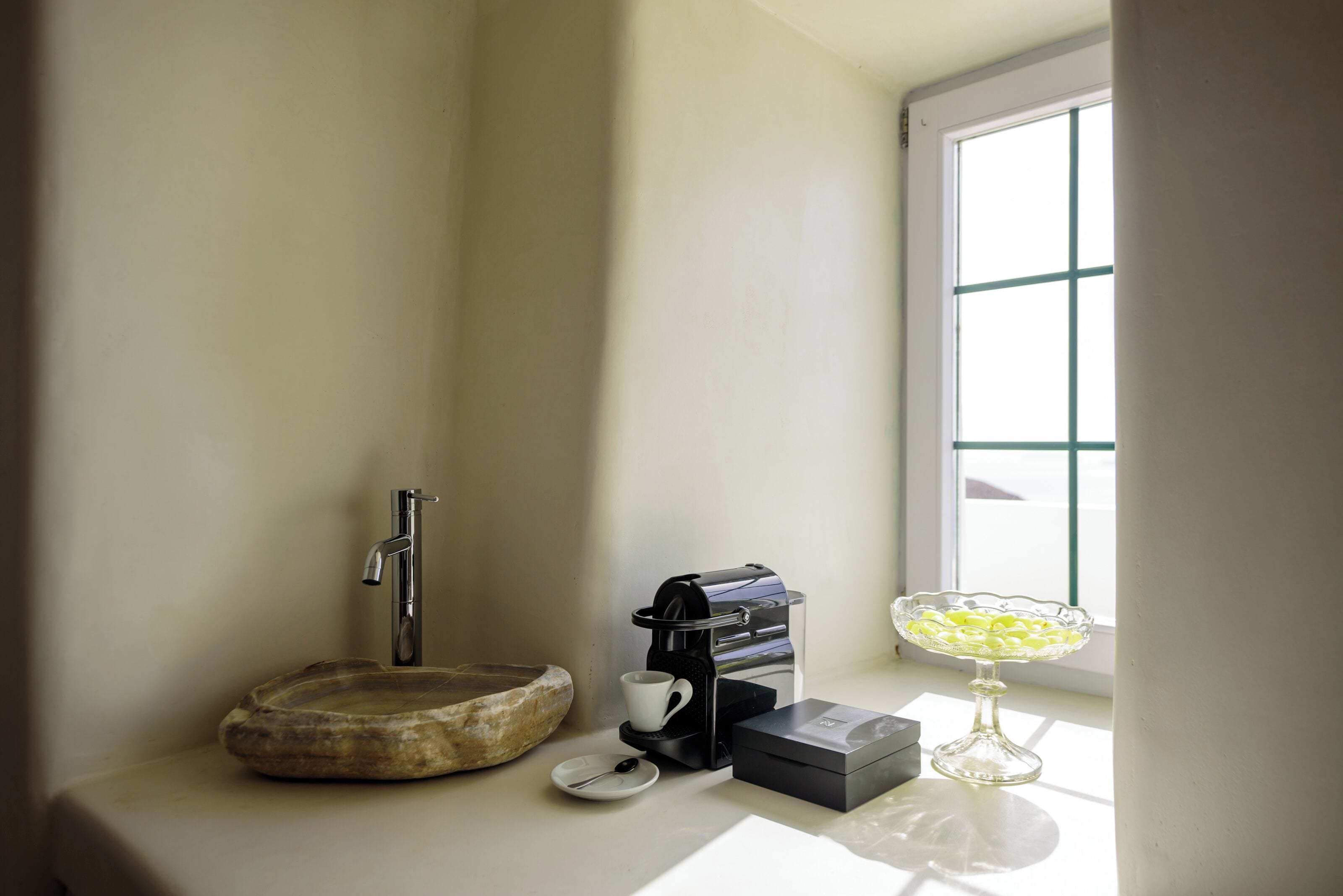 Casa de banho com lavatório de pedra, máquina de café, taça de vidro e janela com persianas.
