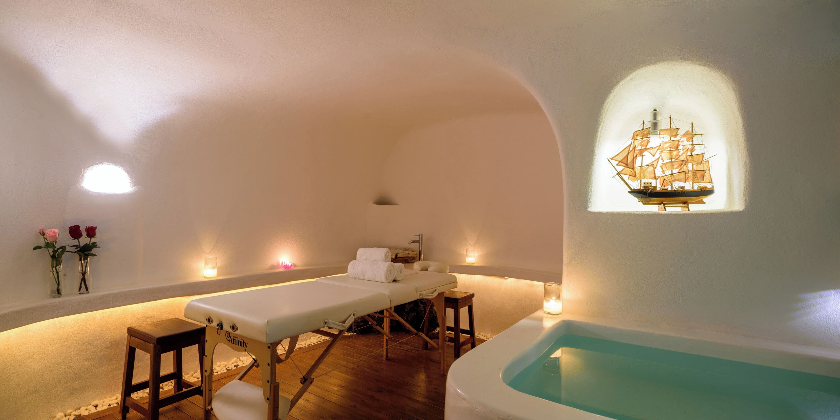 A spa room with a massage table, a small wooden stool, a bathtub, and a wall-mounted ship decoration.