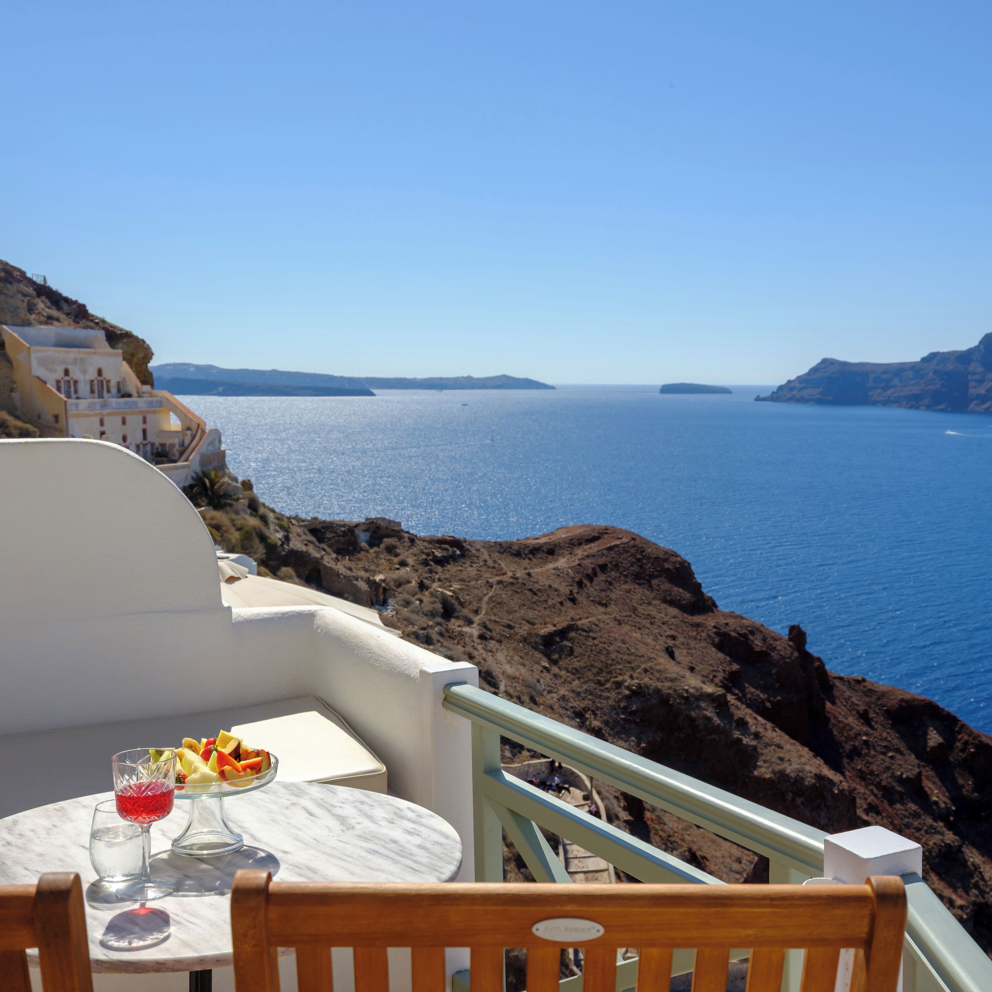 A seaside terrace with a table set for two, overlooking a clear blue sea and rocky coastline.