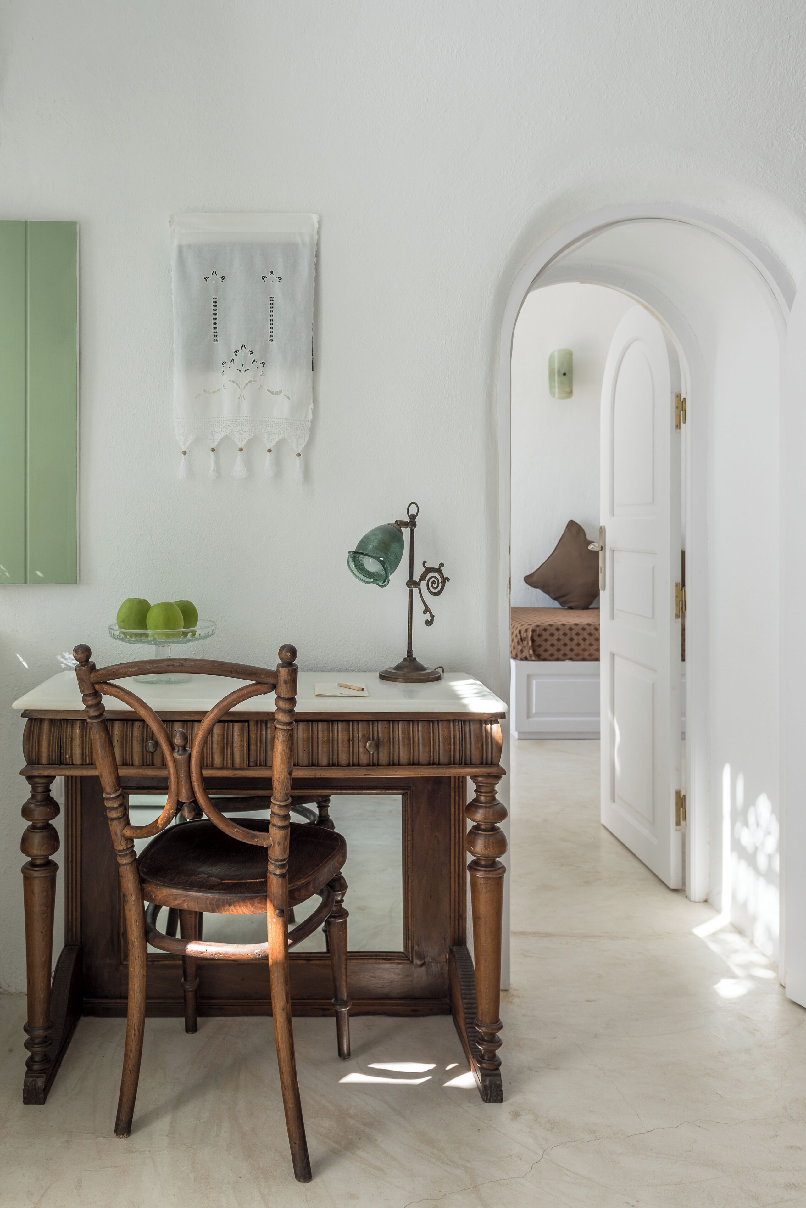 A room with a wooden chair, a desk with a lamp, a bowl of green apples, and a bed with a brown pillow.