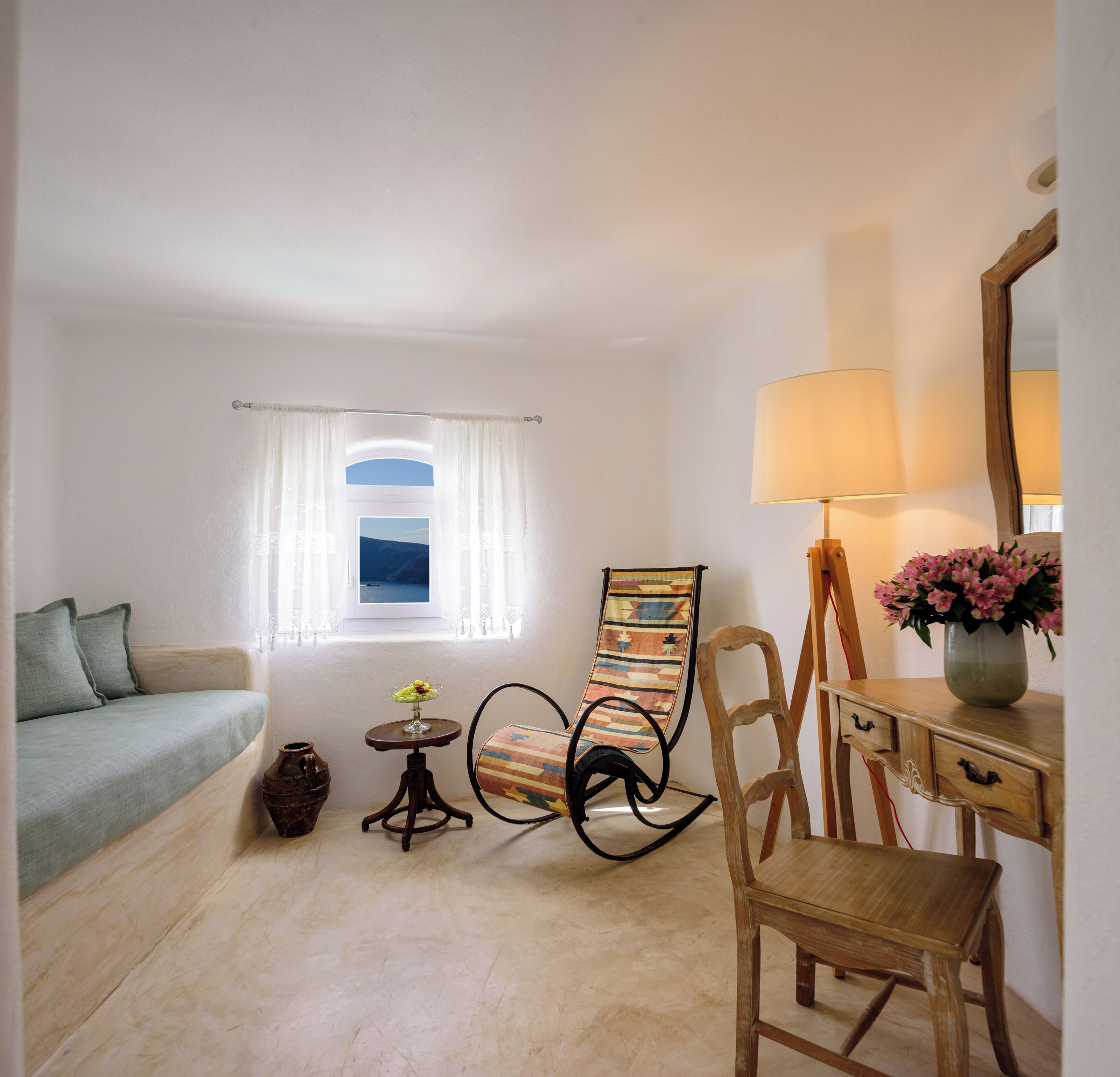 Una habitación acogedora con un sofá, una mesita con un florero, una silla, una lámpara y una ventana con cortinas.