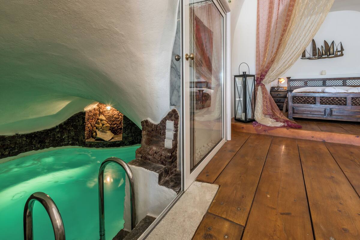 Quarto com hidromassagem em vidro, piso de madeira, cama com cabeceira e um objeto decorativo fixado na parede.