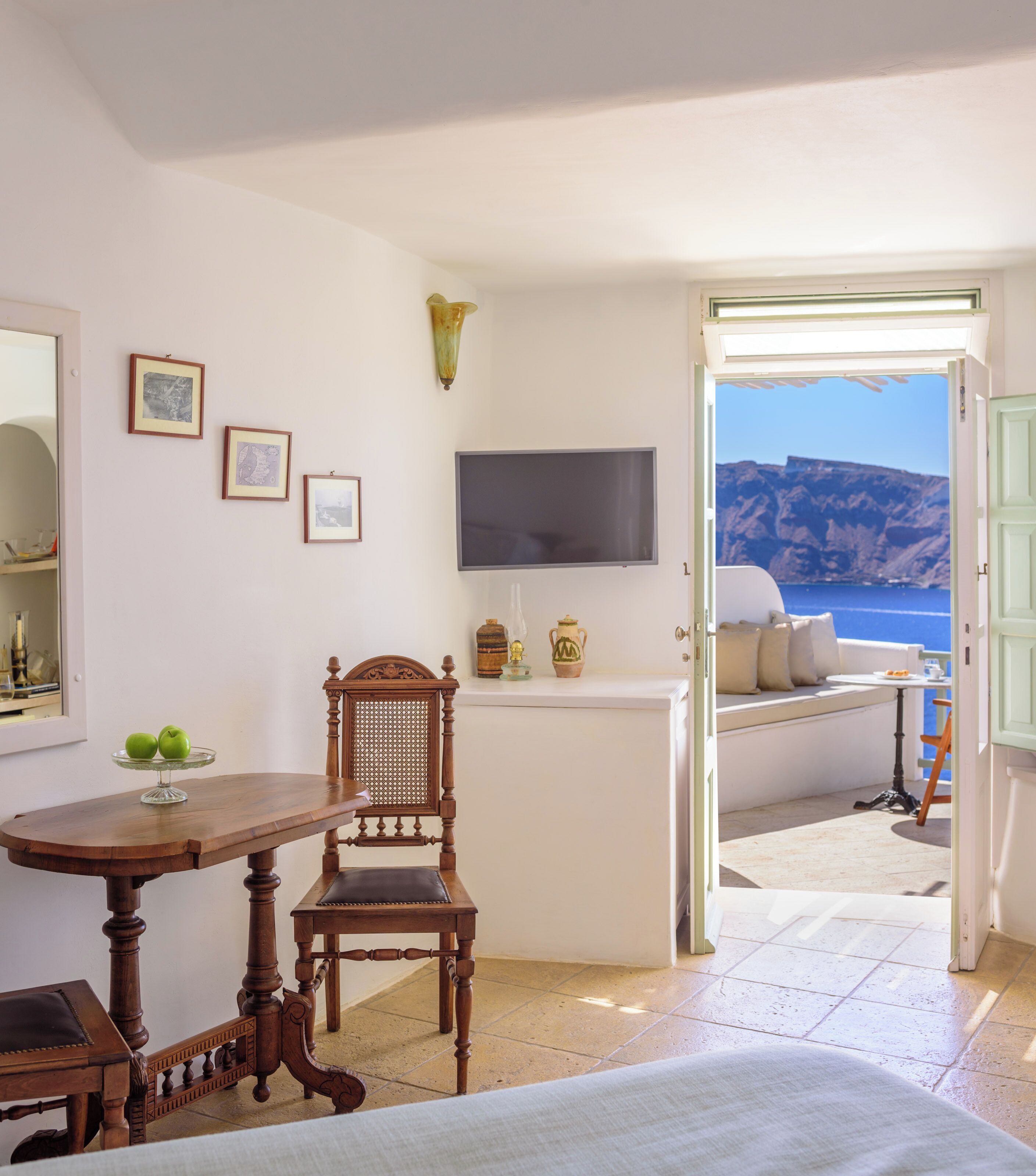 A room with a wooden chair, a small table, a flat-screen TV, and a view of the sea through an open door.