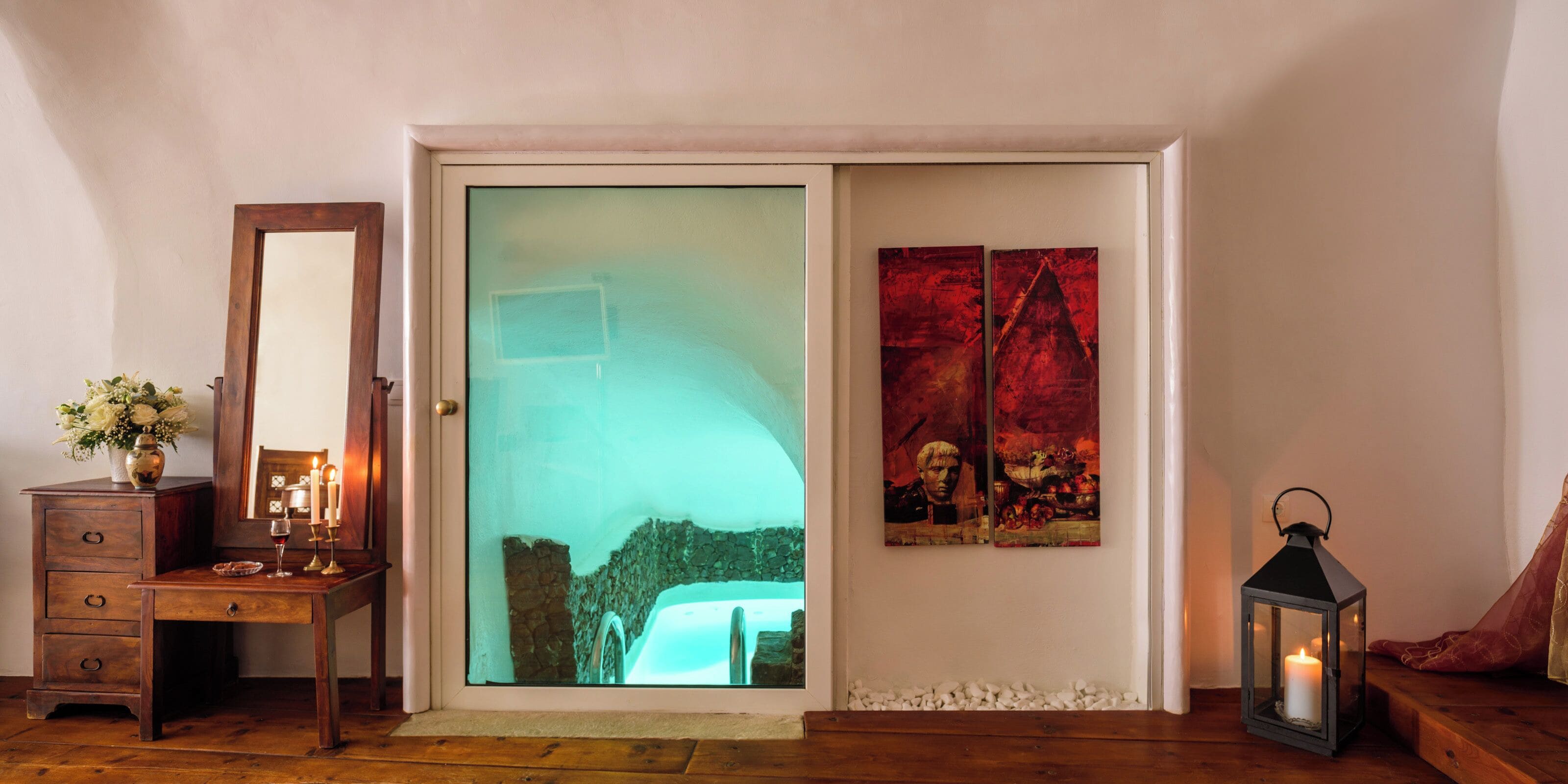 A room with a wooden dresser, a mirror, a glass door with a view of a stone wall, and two paintings on the wall.