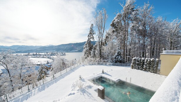 Une piscine extérieure recouverte de neige, avec une eau claire, entourée d’arbres chargés de neige et offrant une vue sur un lac et des montagnes en arrière-plan.