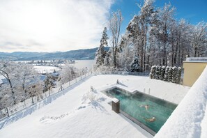 Une piscine extérieure recouverte de neige, avec une eau claire, entourée d’arbres chargés de neige et offrant une vue sur un lac et des montagnes en arrière-plan.