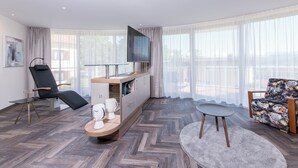 Une chambre d’hôtel moderne dotée d’une grande fenêtre, d’un téléviseur à écran plat, d’un fauteuil noir inclinable, d’une table basse en bois et d’un fauteuil à motifs floraux.