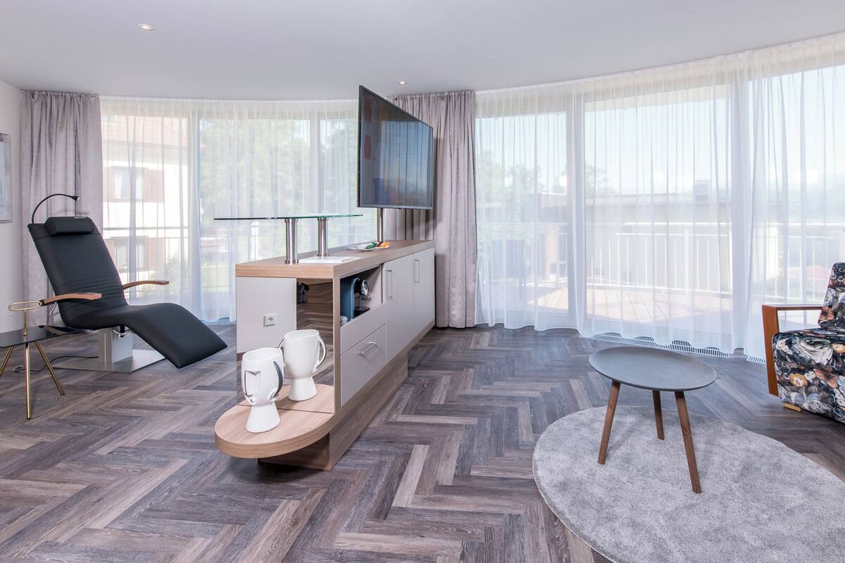 Ett modernt hotellrum med ett stort fönster, en platt-TV, en svart, justerbar fåtölj, ett träbord och en fåtölj med blommönster.