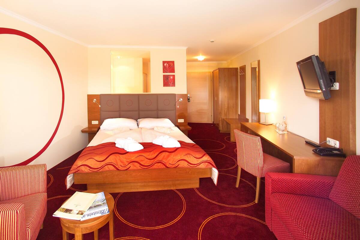 Ett hotellrum med en stor säng, två fåtöljer, ett skrivbord med en dator, en tv och en röd matta.
