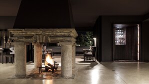 A stone fireplace with a fire, surrounded by pillars and a bench, in a dimly lit room with a view through a window. - Pousada de Manteigas, São Lourenço (Manteigas)
