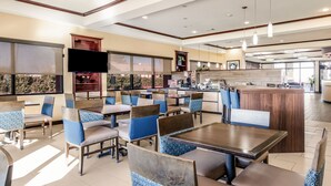 A modern restaurant with multiple dining tables and chairs, a kitchen area, and large windows providing natural light.