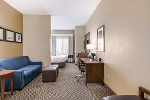 A hotel room with a blue sofa, a small desk with a lamp, a bed, and a view of the outdoors through the window. - Comfort Suites Fort Stockton (Fort Stockton)