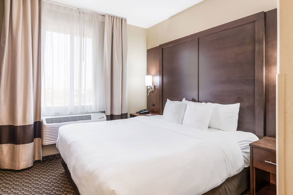 Ein Hotelzimmer mit einem großen Bett, einem Nachttisch, einer Lampe und einem Fenster mit Vorhängen.