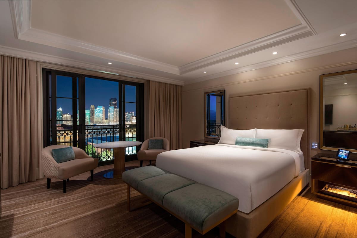 A hotel room with a large bed, a bench, a small table, and a view of the cityscape through large windows.