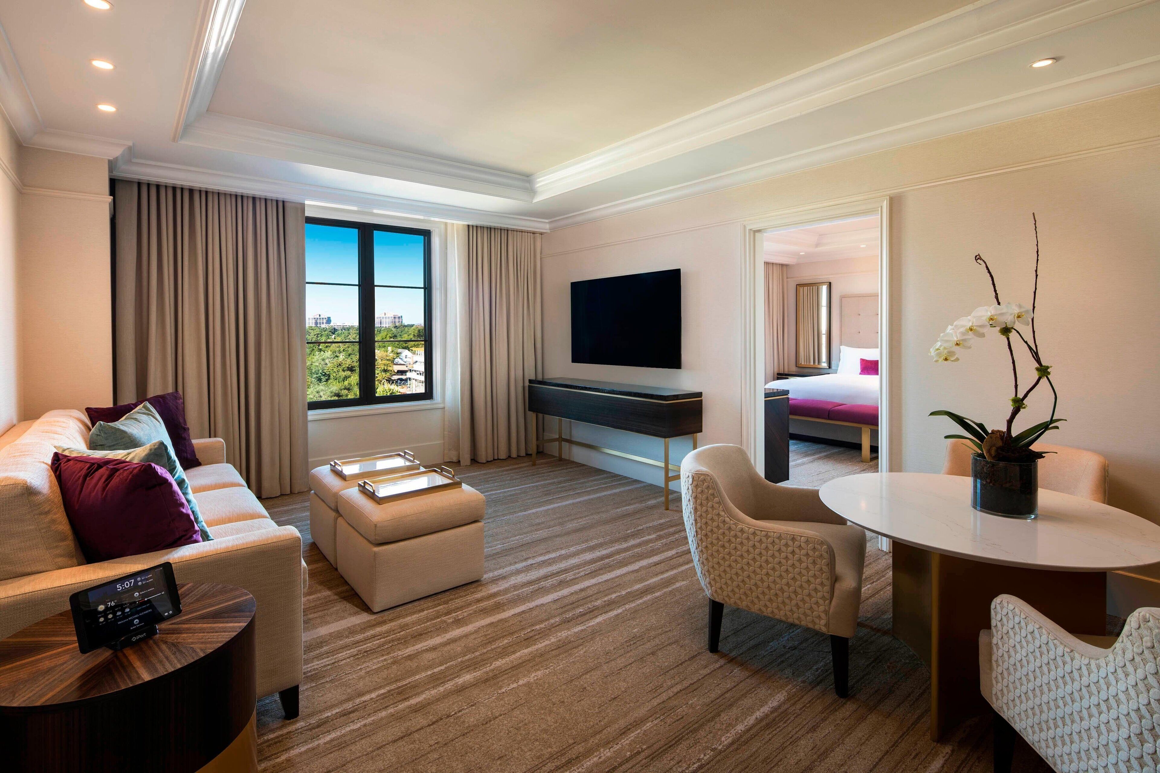 A modern hotel room with a large window, a flat-screen TV, a sofa, a dining table, and a potted orchid.