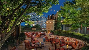 An outdoor seating area with multiple tables and chairs, surrounded by greenery and a fire feature.