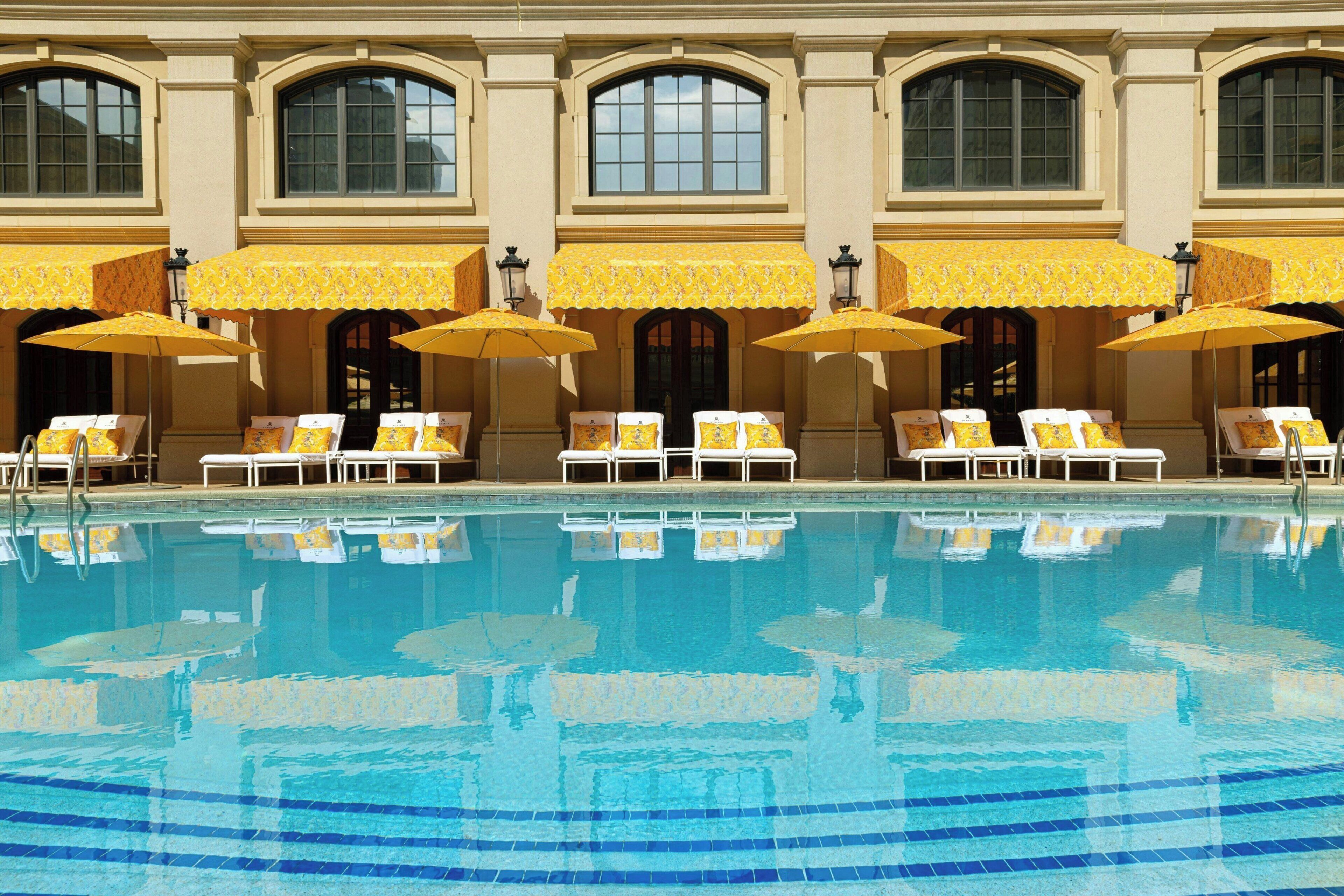 A pool with lounge chairs and umbrellas, a building with arched windows, and a tiled pool deck.