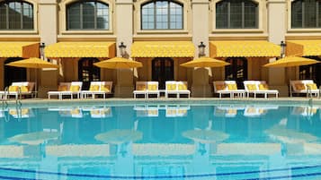 A pool with lounge chairs and umbrellas, a building with arched windows, and a tiled pool deck.