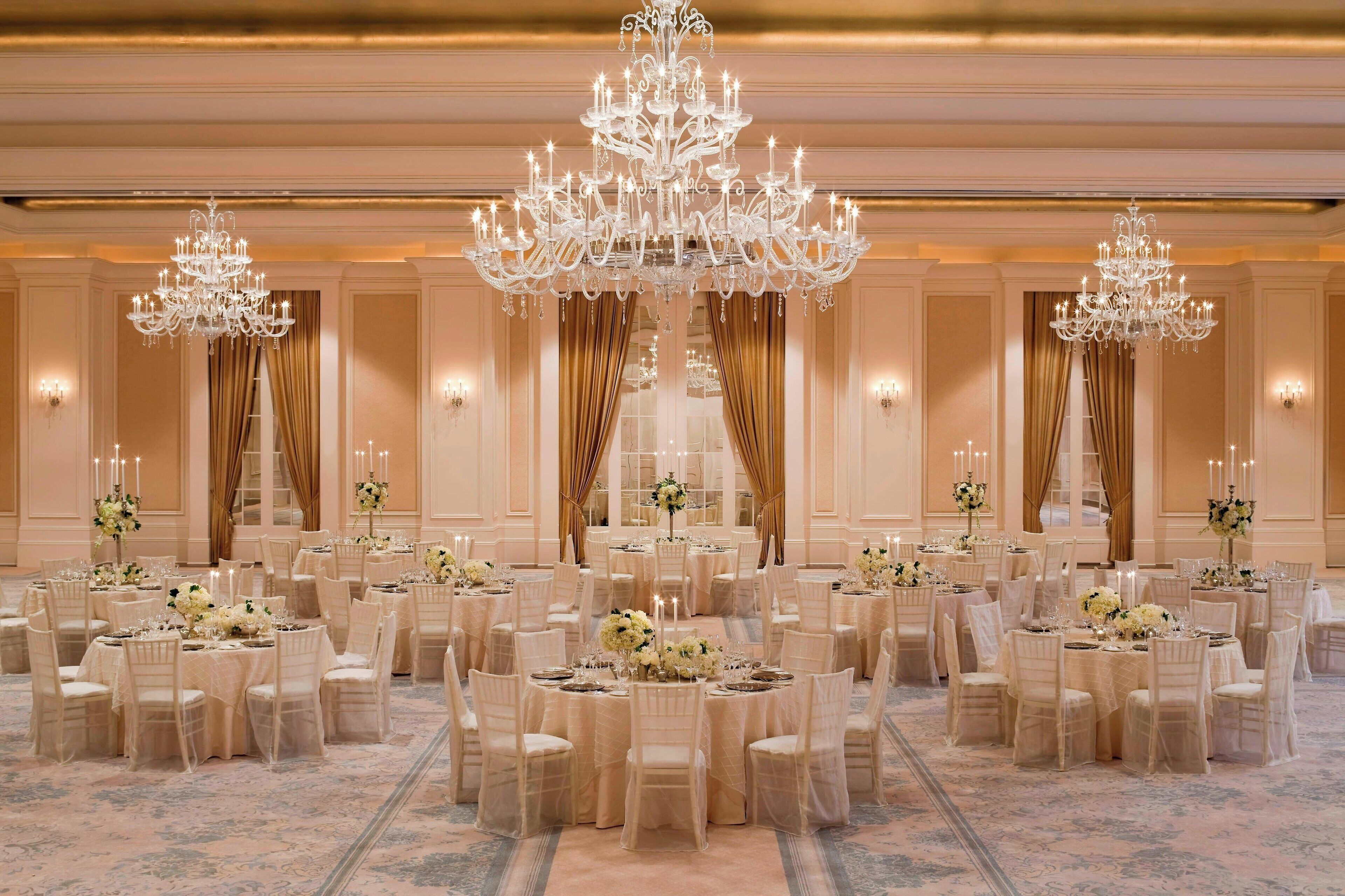 Un salón de banquetes con mesas redondas, sillas blancas, candelabros y centros de mesa florales.