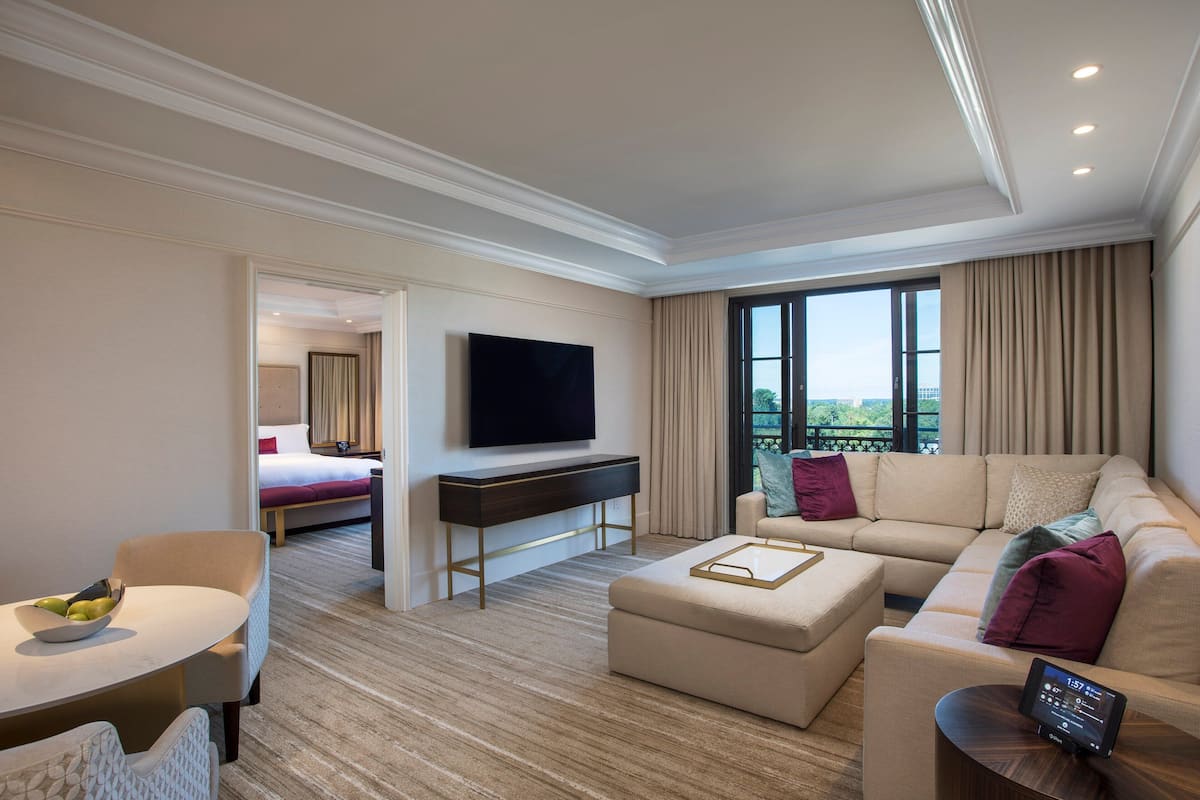 A modern hotel room with a large sofa, a dining table, a flat-screen TV, and a balcony with a view.