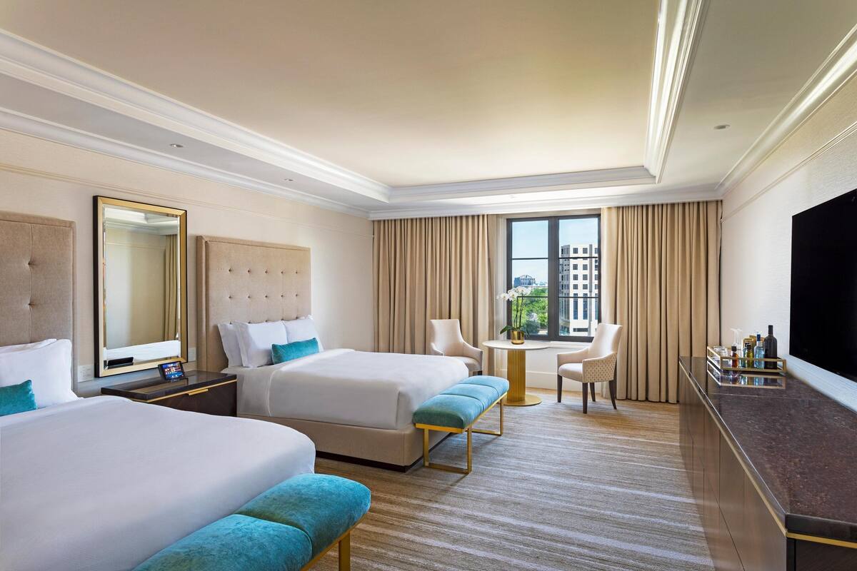 A hotel room with two beds, a TV, a small table, and a view of the city.