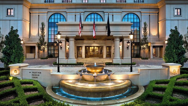 A grand hotel facade with a central fountain, flagpoles, and well-manicured gardens.