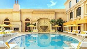 A pool area with lounge chairs and umbrellas, a fountain, and a building facade with arches and columns.
