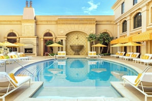 Un espace piscine dotĂ© de chaises longues et de parasols, dâune fontaine et dâune façade ornĂ©e dâarches et de colonnes.