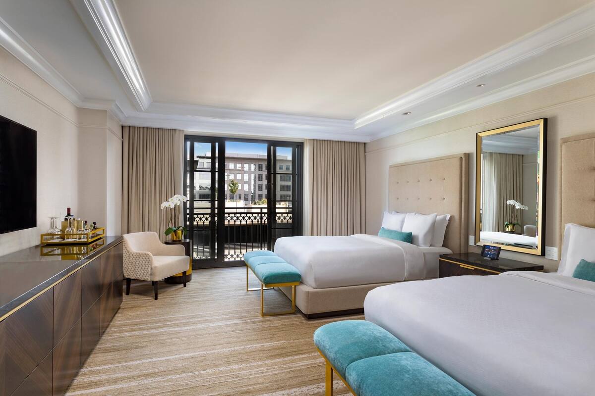 A hotel room with a large bed, a smaller bed, a sofa, a desk with a flat-screen TV, and a balcony with a view of buildings.