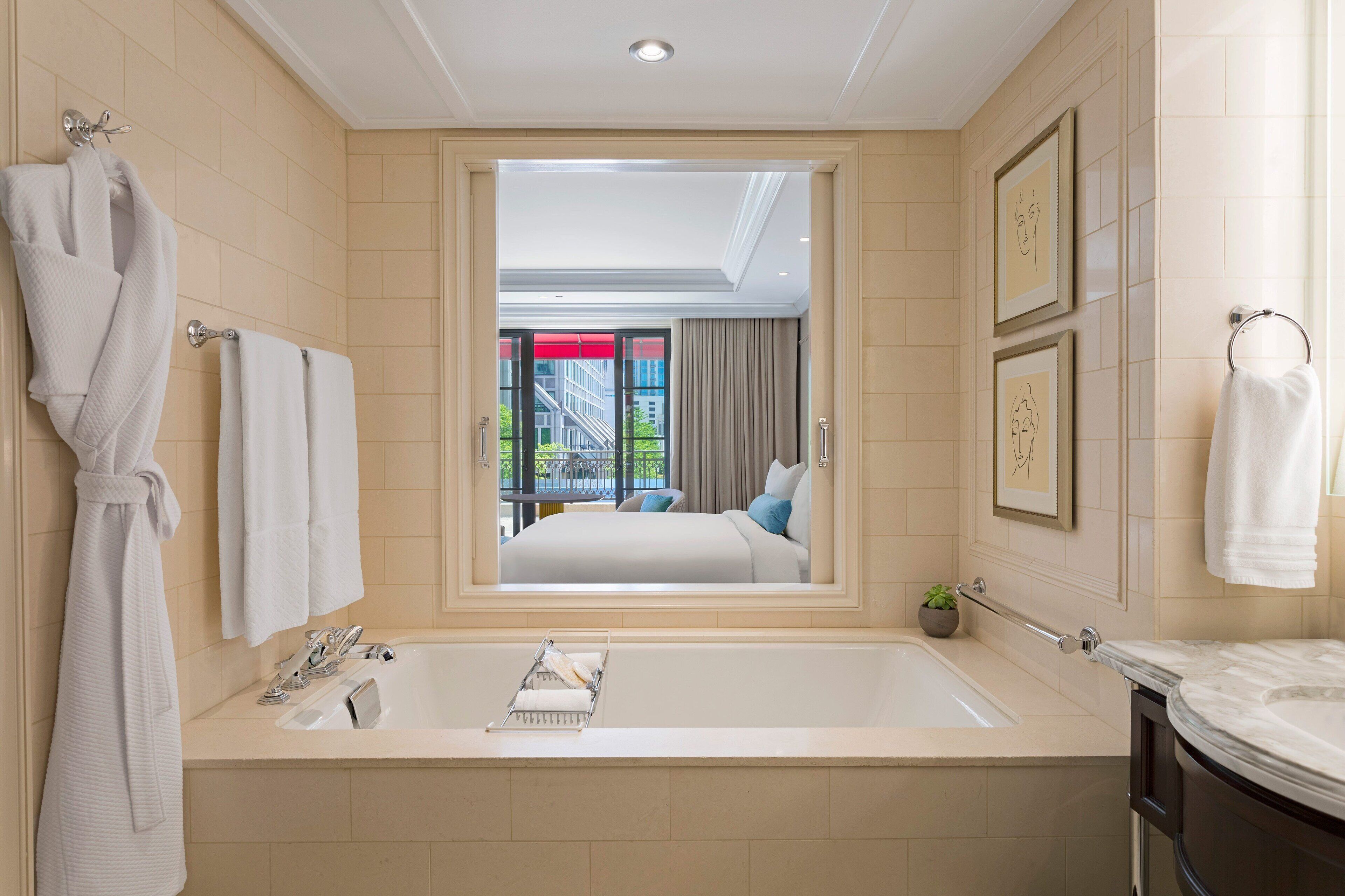A modern bathroom with a large bathtub, a mirror reflecting a bedroom, and a view of a building outside.