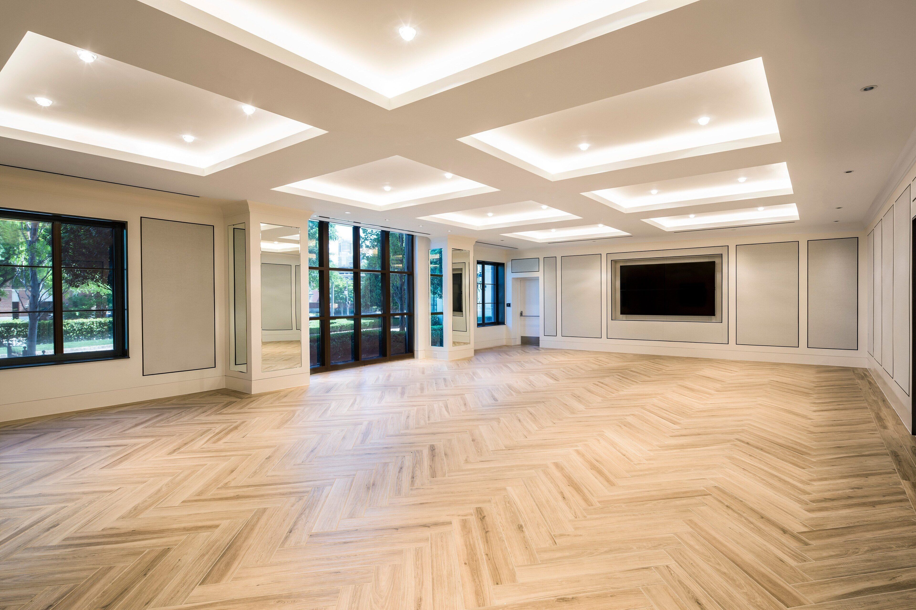 A spacious room with a herringbone floor pattern, recessed ceiling lighting, large windows, and a flat-screen TV mounted on the wall.