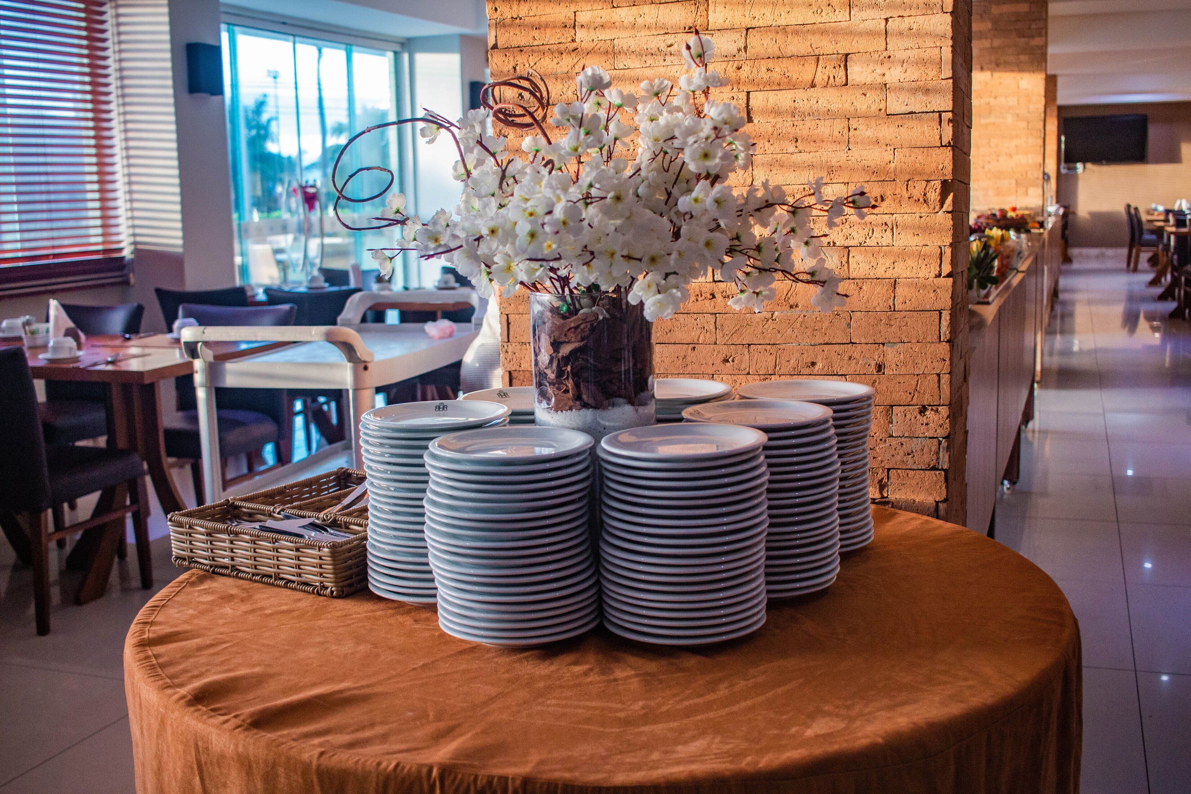 Un comedor con pared de ladrillo, una mesa con platos apilados y un jarrón con flores blancas.