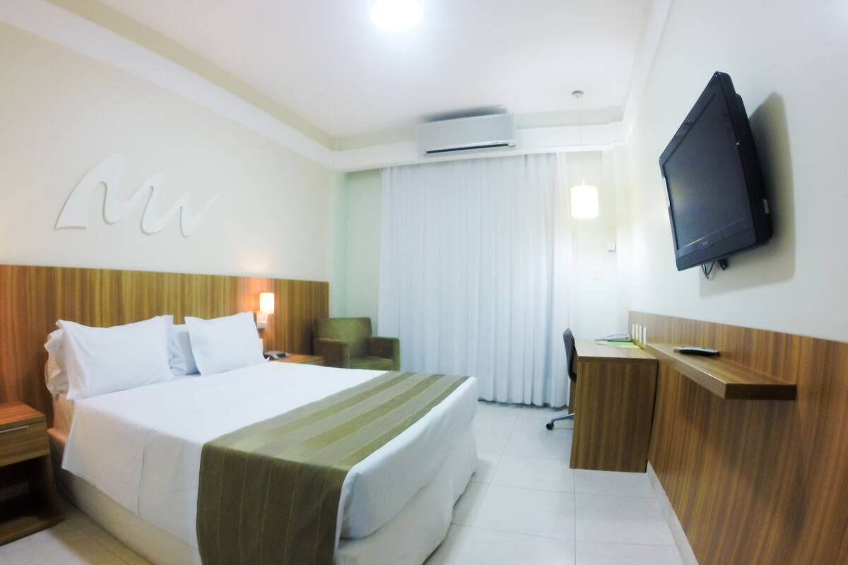 A hotel room with a bed, a desk, a chair, a television, and an air conditioning unit.