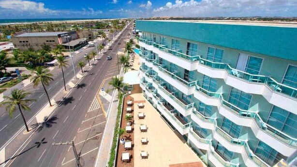 A multi-story hotel with a pool, a street with palm trees, and a beach in the background. - Real Classic Hotel (Aracaju)