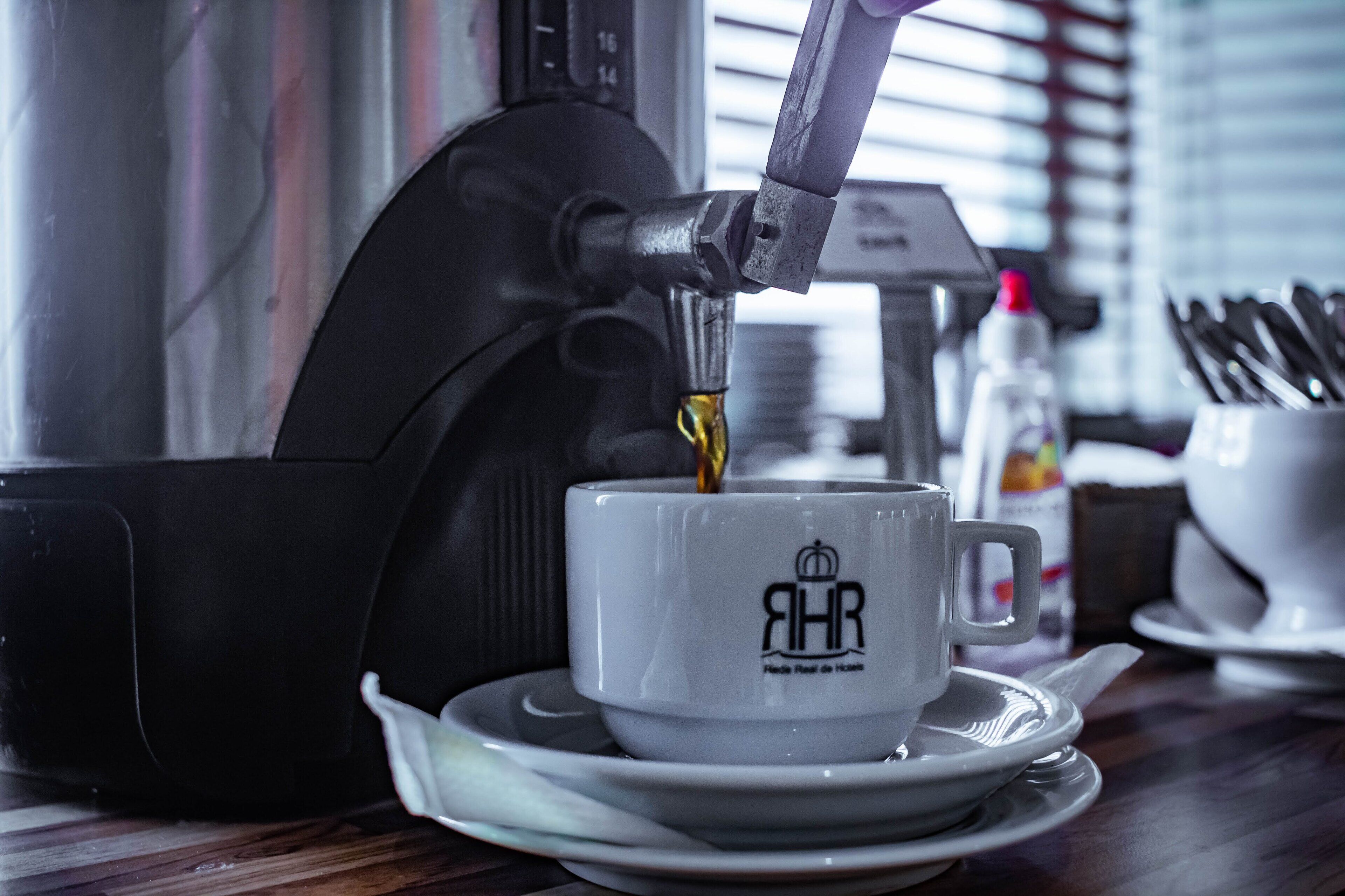 Eine Kaffeemaschine füllt Kaffee in eine Tasse mit einem Logo, eine Gewürzflasche und ein Set von Besteck liegen auf einem Tisch.