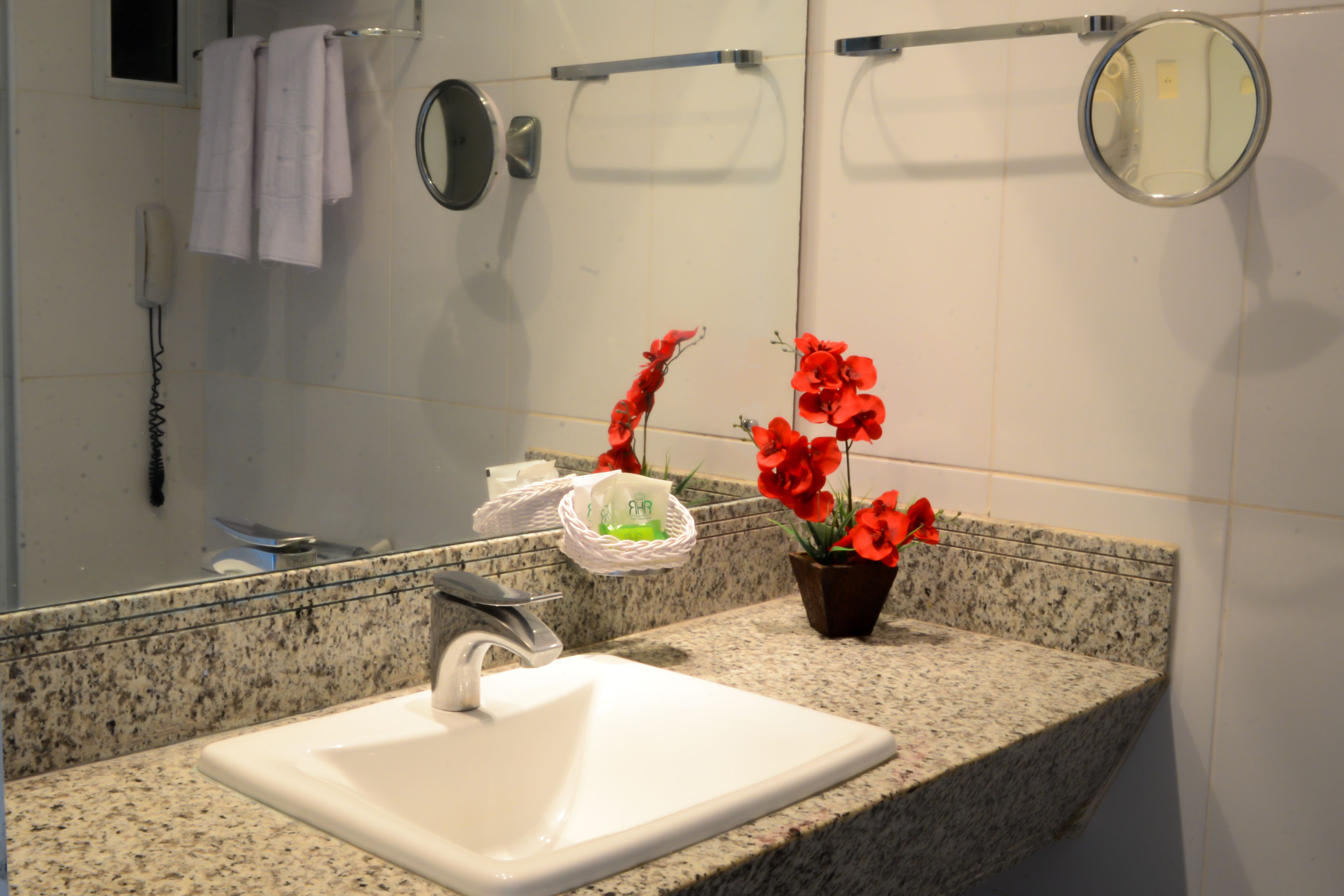 Un baño con lavamanos blanco, encimera de granito, un espejo, perchero para toallas, teléfono y una pequeña maceta con flores rojas.