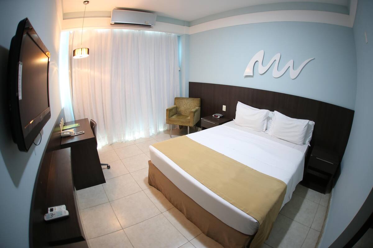 A hotel room with a bed, a desk, a chair, a TV, and a wall-mounted artwork.