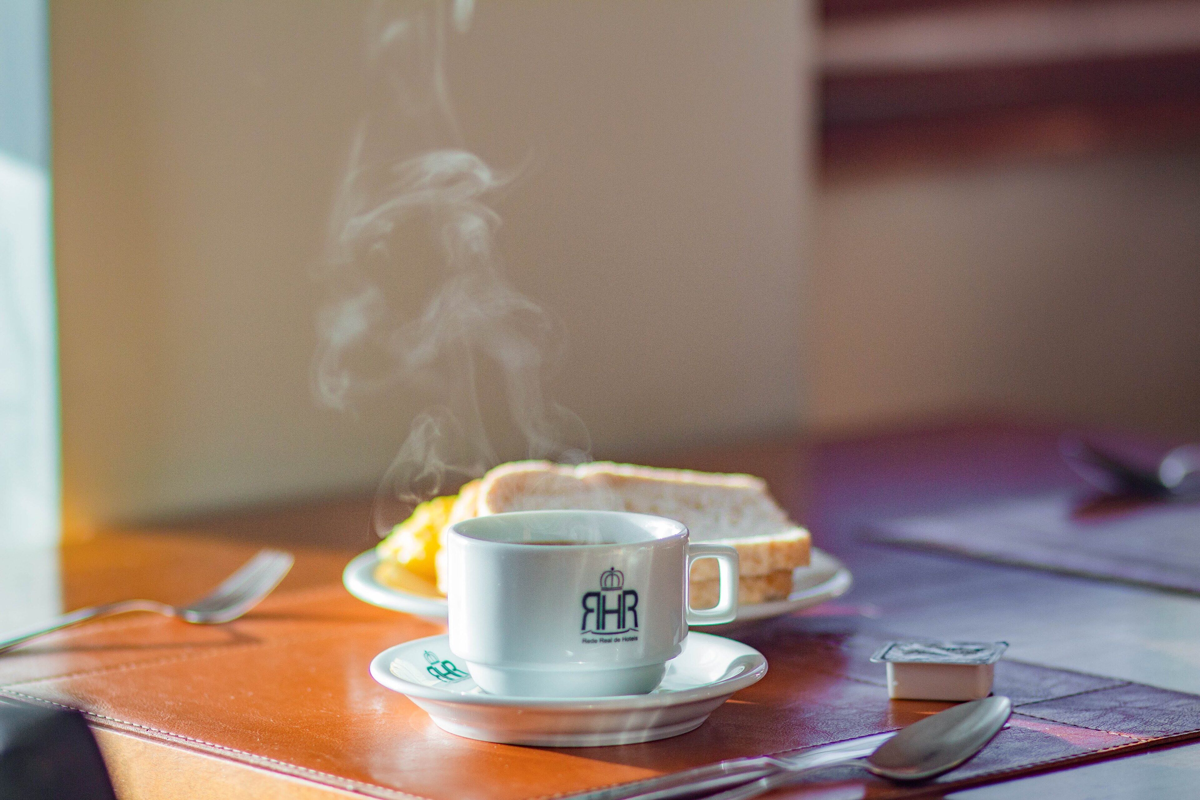 Een kop koffie op een schotel met stoom, een snee toast en een klein bakje boter op een houten tafel.