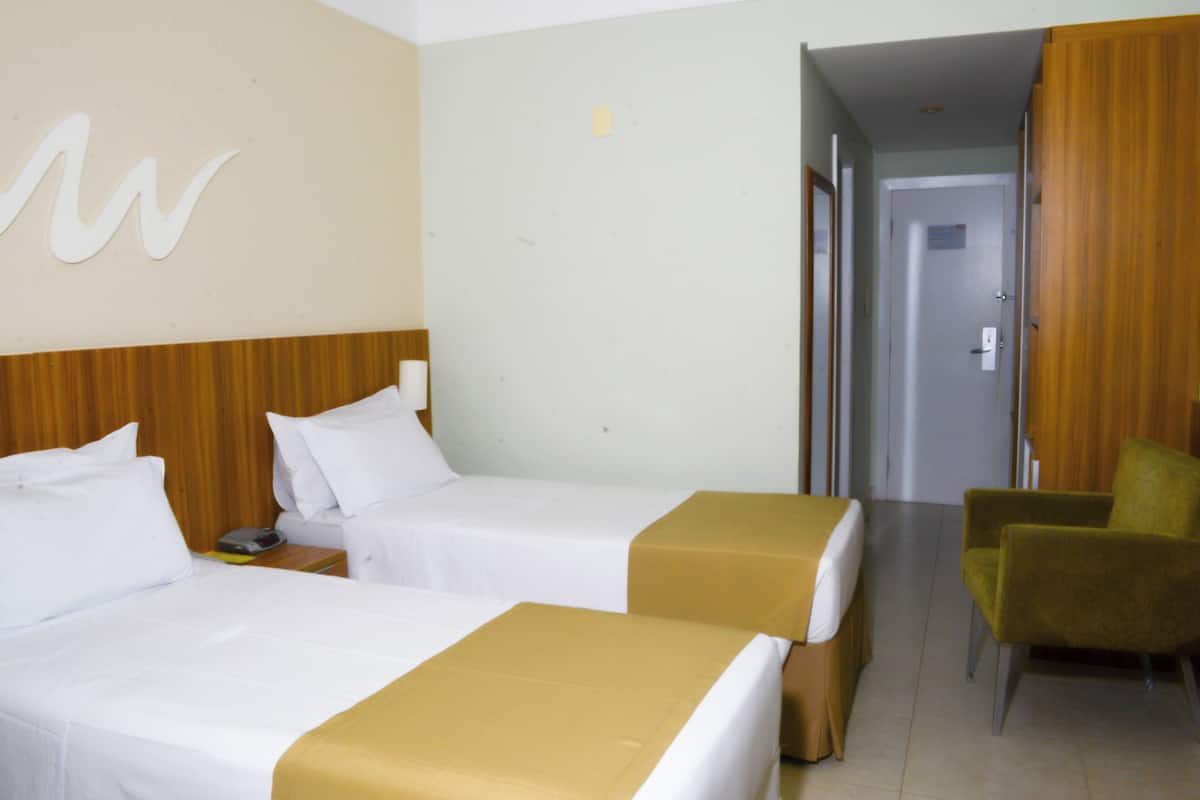 A hotel room with two beds, a wooden headboard, a green armchair, and a door leading to another room.