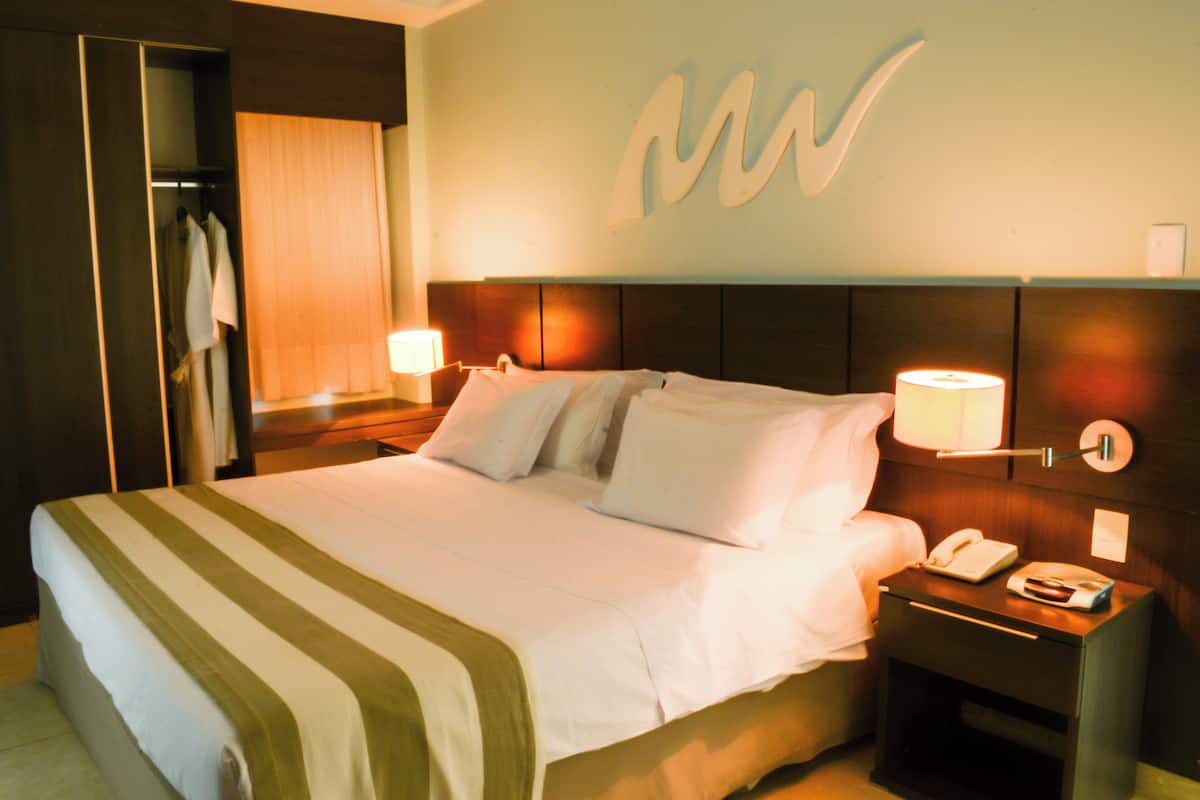 A hotel room with a large bed, bedside lamps, a nightstand with a phone and a remote control, and a mirror with a towel hanging on it.