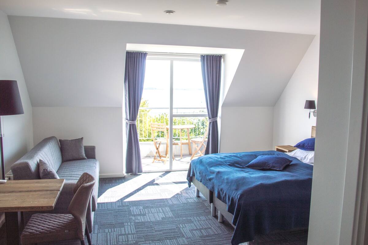 A bedroom with a bed, a sofa, a table, and a chair. There is a balcony with a table and chairs.