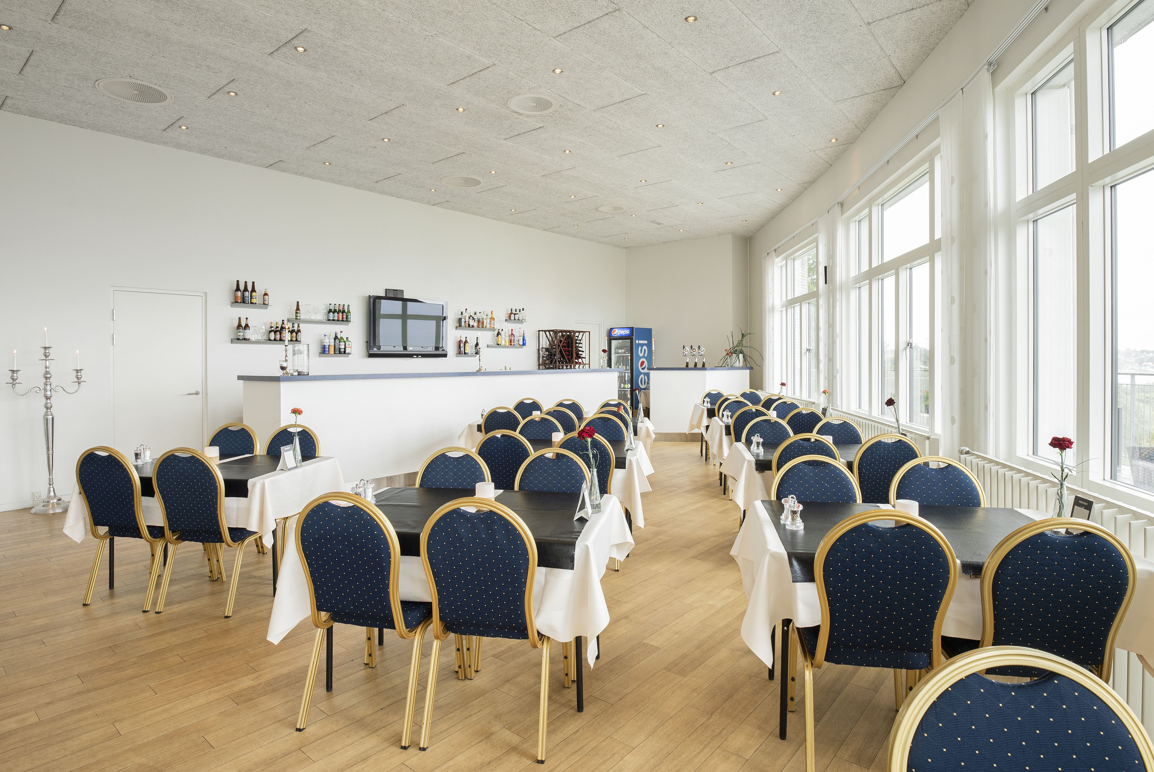 A spacious conference room with long tables and chairs, a bar area with bottles, and large windows providing natural light.